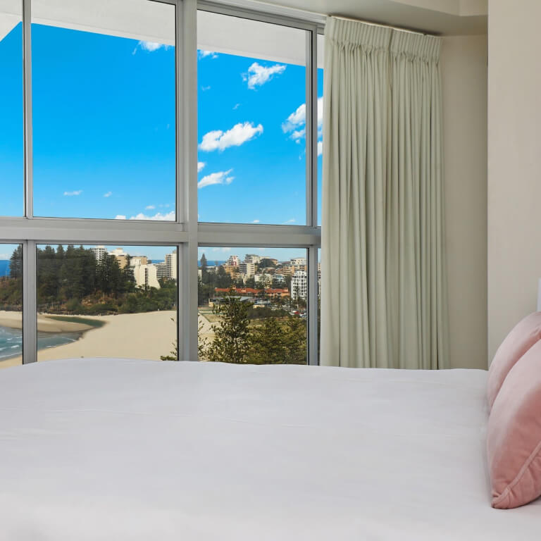 Bedroom with large windows at ULTIQA Points North Coolangatta offering panoramic ocean and coastline views, featuring a neatly made bed with soft pink cushions.