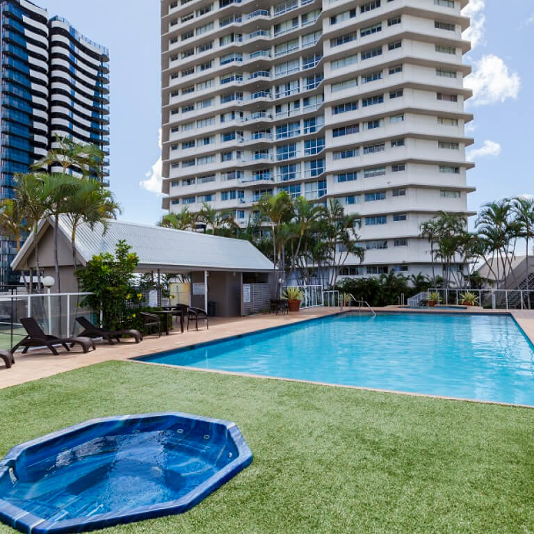 Outdoor swimming pool and spa area at ULTIQA Points North Coolangatta, surrounded by sun loungers, palm trees, and high-rise buildings under a bright blue sky.