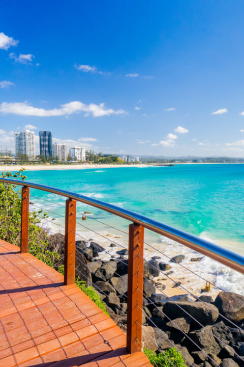 Scenic oceanfront boardwalk in Coolangatta with turquoise water, golden sand, and high-rise buildings along the coastline under a bright blue sky.