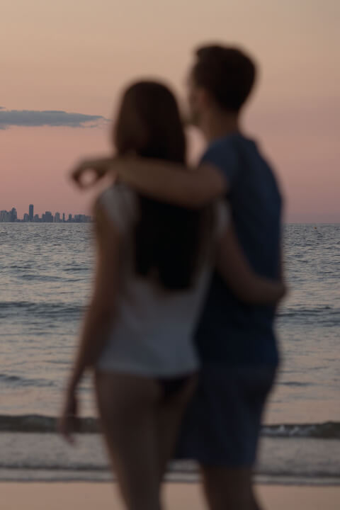 Couple standing on the beach at sunset, admiring the Gold Coast skyline across the water.