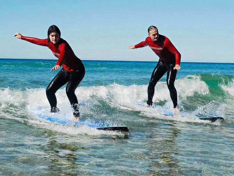 Two people learning to surf, balancing on small waves during a lesson at the beach.