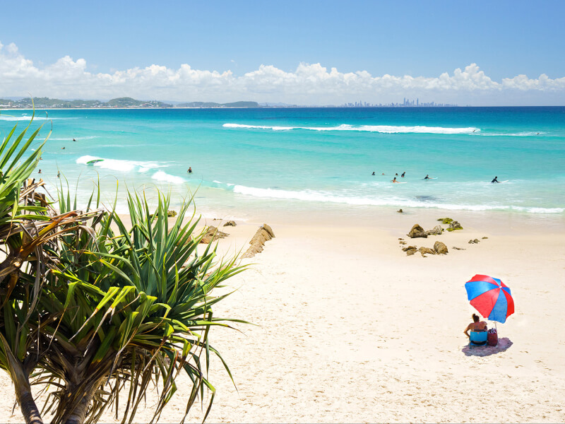 Scenic Coolangatta Beach with turquoise water, surfers, and a couple under a red and blue umbrella.