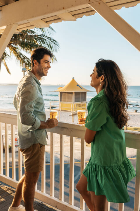 Couple enjoying drinks on a beachfront balcony overlooking the ocean and a yellow lifeguard tower at sunset.