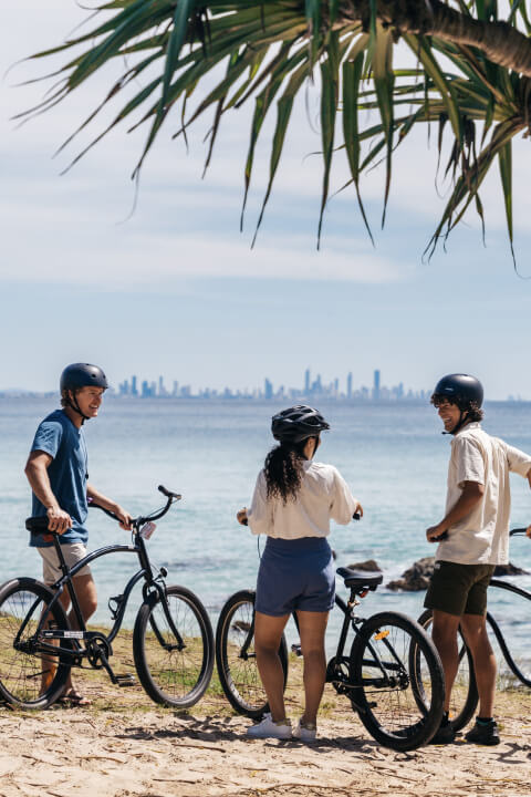 Three friends with bikes stop to enjoy the ocean view along the coastline, with the Gold Coast skyline visible in the distance.