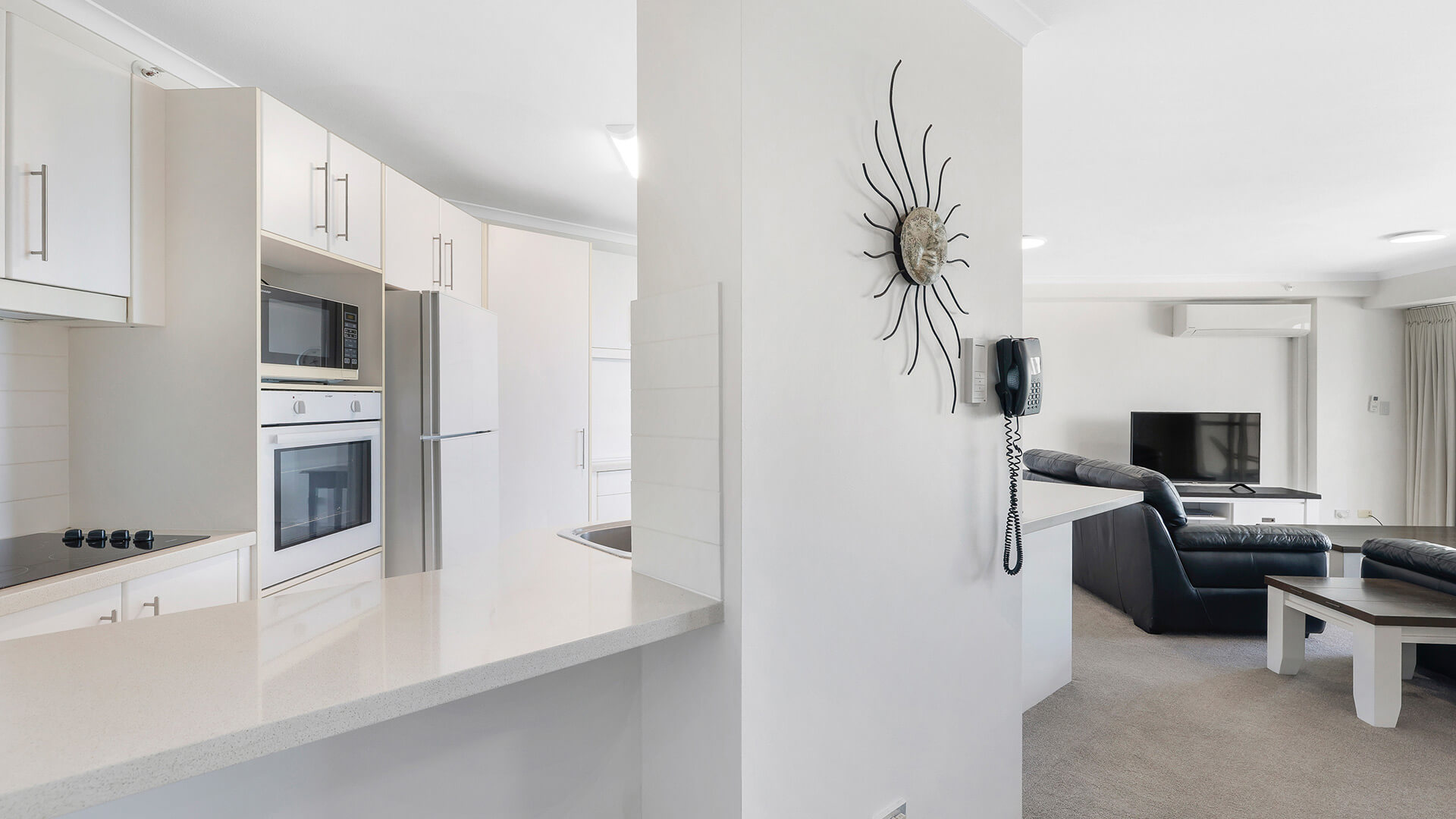 Open-plan layout showing a modern white kitchen with built-in appliances & breakfast bar, looking into the living area with leather seating and TV.