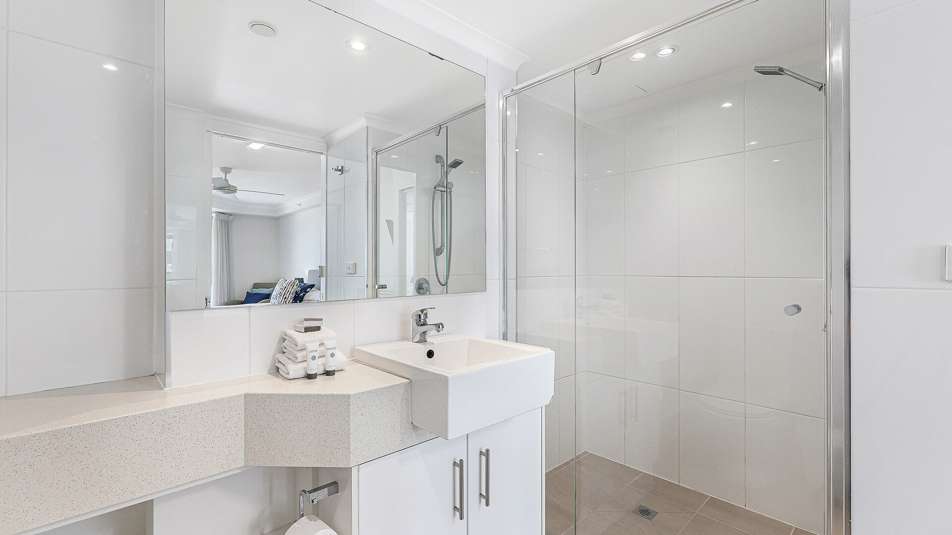 Bright modern bathroom with a large mirror, white vanity, glass-enclosed walk-in shower, and neatly folded towels on the counter.