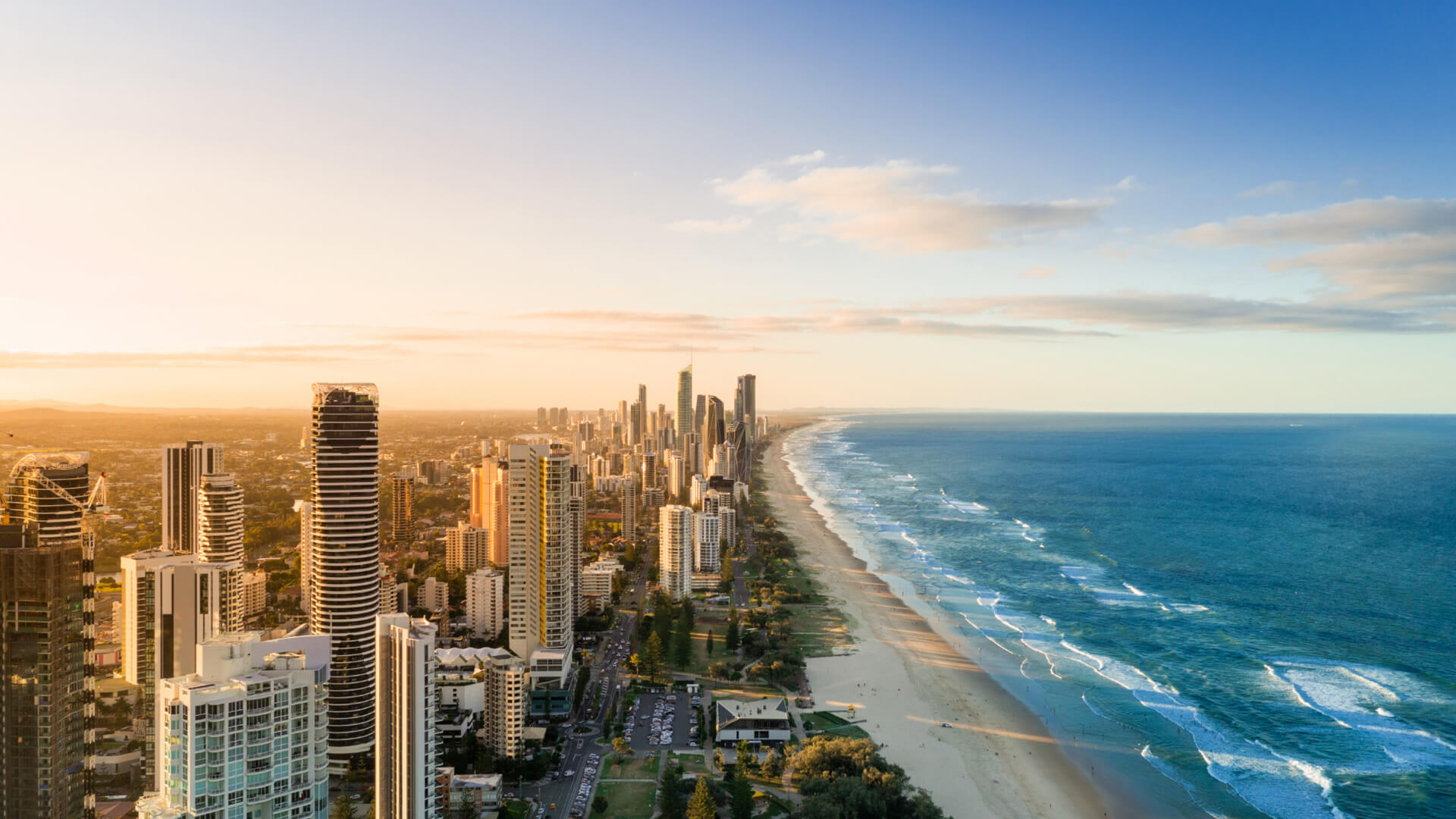 Aerial view of the Gold Coast skyline and beachfront at sunset with golden light over the city and waves along the shore.